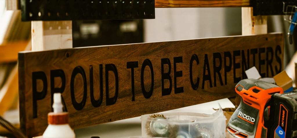 Carved wooden sign reading “Proud to Be Carpenters” behind a Ridgid sander on a workbench